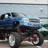 A custom lifted blue pickup truck with oversized wheels and red suspension components, shown in a social media post. The truck is parked outdoors in front of a modern building.